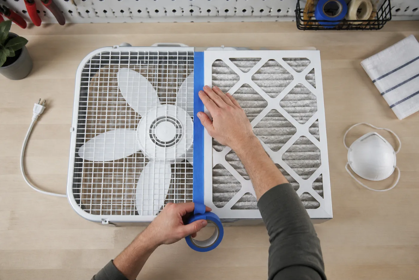 Hands securing a high-efficiency filter to an unplugged box fan for temporary wildfire-smoke filtration